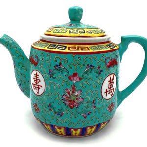 Könnte beinhalten: Türkisgrüne Keramik-Teekanne mit Deckel, Henkel und Ausguss. Die Teekanne ist mit floralen Mustern, chinesischen Schriftzeichen und roten, gelben und blauen Akzenten verziert. Der Deckel hat einen kleinen Knopf.