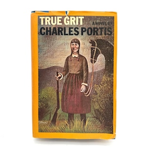 May include: A hardback book titled True Grit by Charles Portis with a yellow border. The cover features an illustration of a person with dark hair holding a rifle, standing next to a dark horse in a landscape with rolling hills and green grass.