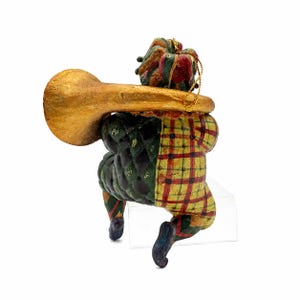 May include: A jester figurine ornament wearing a multi-colored hat and a suit with a green quilted pattern on one side and a yellow and red plaid pattern on the other. The jester holds a large, textured gold-colored trumpet and has a gold string.