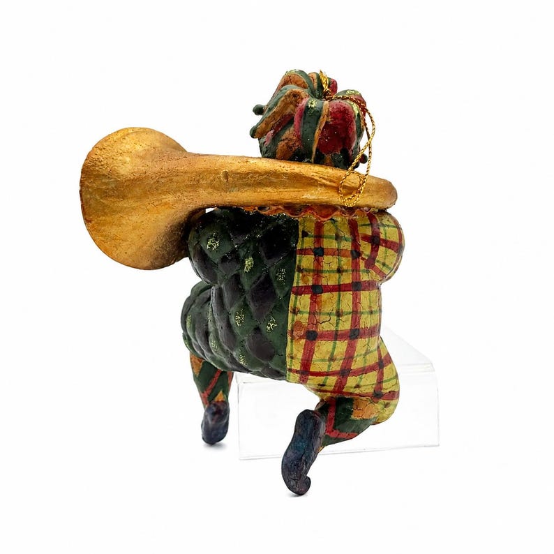 May include: A jester figurine ornament wearing a multi-colored hat and a suit with a green quilted pattern on one side and a yellow and red plaid pattern on the other. The jester holds a large, textured gold-colored trumpet and has a gold string.