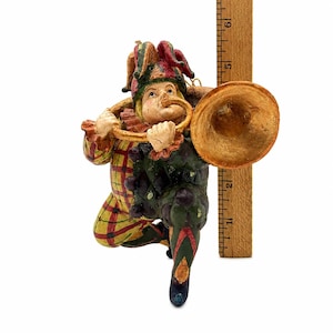 May include: A jester figurine wearing a red, yellow, and green patterned outfit and a multi-pointed hat, playing a gold-colored trumpet. The figure is positioned next to a wooden ruler, showing a height of approximately 6 inches from base to top.