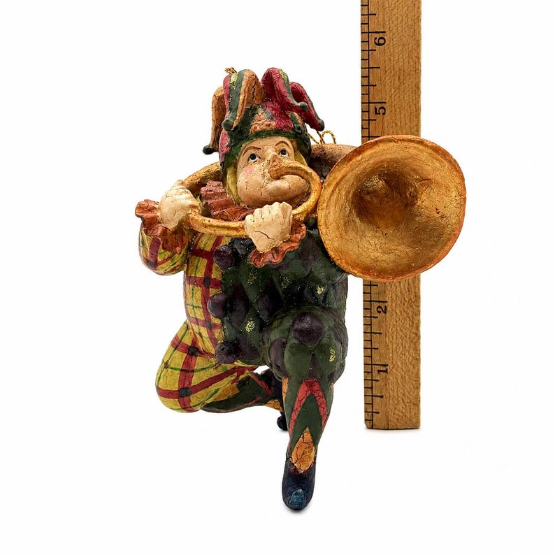 May include: A jester figurine wearing a red, yellow, and green patterned outfit and a multi-pointed hat, playing a gold-colored trumpet. The figure is positioned next to a wooden ruler, showing a height of approximately 6 inches from base to top.