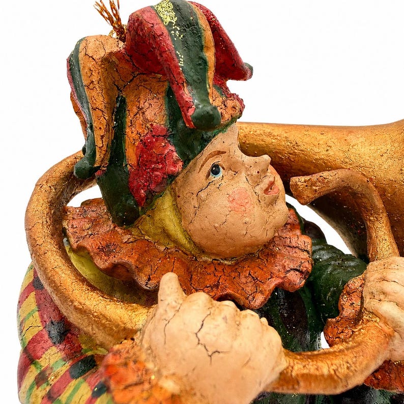 May include: A decorative figurine of a jester wearing a multi-colored hat with red, green, and yellow sections. The jester has a cracked, textured finish, a ruffled collar, and is holding a large, curved, gold-colored musical instrument.
