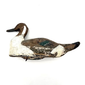 May include: A weathered wooden duck decoy featuring a brown head, a white chest and neck, and a brown body with a distinct green patch on the wing area. The paint shows signs of age and wear, with a dark beak and a pointed tail section.