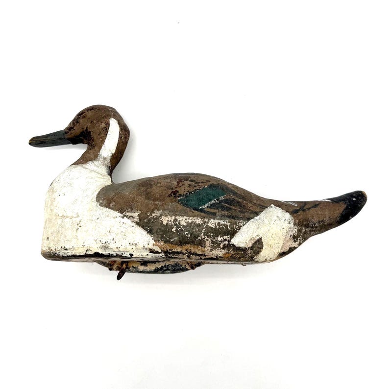 May include: A weathered wooden duck decoy featuring a brown head, a white chest and neck, and a brown body with a distinct green patch on the wing area. The paint shows signs of age and wear, with a dark beak and a pointed tail section.