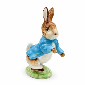 May include: A ceramic figurine of a brown and white rabbit standing on a green base. The rabbit is wearing a bright blue jacket with small orange buttons. The figure has long ears, dark eyes, and a glossy finish, captured in a standing pose.
