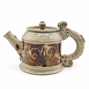 May include: A ceramic teapot with a light gray glaze and a central band of dark brown, swirling patterns. The handle is sculpted into a curved, segmented form with decorative scrollwork at the top and bottom, and a matching lid sits on top.