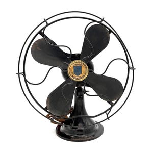 May include: A vintage black electric desk fan with four metal blades and a protective wire cage. The center of the fan features a circular gold and blue emblem with the text GrayBar and Western Electric printed clearly on the front surface.