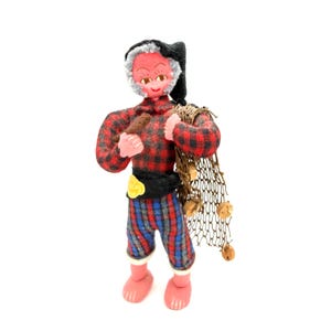 Op de afbeelding: Handgemaakte pop van een visser, gekleed in een rood-zwart geruit overhemd, blauw-rood gestreepte broek en een zwarte hoed. De pop draagt een visnet en een klein bruin voorwerp.