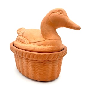May include: A terracotta duck-shaped container with a basket-weave base. The duck's head and body form the lid, while the basket-like base serves as the container. The container is a warm, earthy orange color.