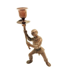 May include: A bronze-colored candlestick holder in the shape of a monkey holding a torch. The monkey is wearing a detailed outfit and has a serious expression. The candle holder is a small, decorative item.