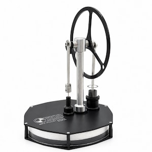 May include: A Stirling engine model featuring a black hexagonal base with the text American Stirling Company MM-7 Stirling Engine, a central silver vertical pillar, a black flywheel, and silver mechanical linkages on a white background.