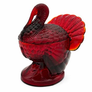 May include: A translucent ruby red glass candy dish shaped like a turkey. The lidded container features intricate molded feather textures on the body, wings, and fanned tail, with the turkey head forming the handle of the lid atop a circular base.