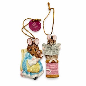 May include: Two Beatrix Potter Hunca Munca hanging ornaments with gold strings. One depicts a mouse in a blue dress holding a baby mouse. The other shows a mouse with glasses reading a newspaper while sitting on a pink thread spool. Both feature a round purple tag.