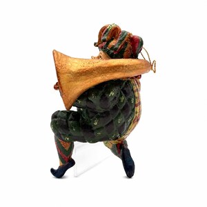 May include: A jester ornament wearing a green and black checkered suit with a matching hat, playing a large gold-colored trumpet. The figure features red and green striped leggings and dark blue curled shoes, with a gold hanging cord attached.