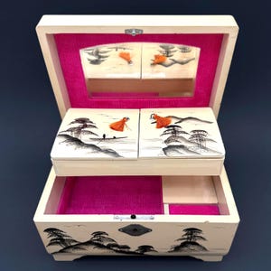May include: Open, vintage jewelry box with a cream-colored exterior and a pink velvet interior. The lid and drawer fronts feature painted landscape scenes with orange accents. A small mirror is visible inside the lid.