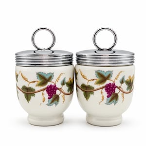 May include: Two cream-coloured ceramic egg coddlers with a grape vine pattern featuring purple grapes and green leaves. Each coddler has a polished silver-tone metal lid with a circular ring handle on top and a ribbed edge design.