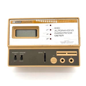 May include: A beige and gray 830 autoranging capacitance meter with a digital display. The meter has a power range switch, a zero adjustment, and input jacks. The text "BK Precision Dynascan Corporation" is visible.