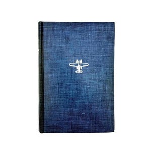 May include: A hardcover book with a textured blue cover. A white airplane graphic is centered on the front. The spine is black. The book is set against a white background.