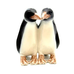 May include: A ceramic figurine of two penguins, standing close together. The penguins have black backs, white bellies, and brown beaks and feet. The penguins are touching beaks, suggesting affection. The figurine is on a white background.