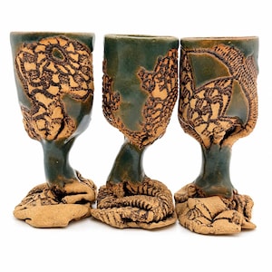 May include: Three ceramic goblets featuring a dark green glaze and textured, tan-coloured clay accents. Each vessel has a stem that transitions into a wide, organic, and uneven base with intricate, raised, and dark-toned surface patterns.