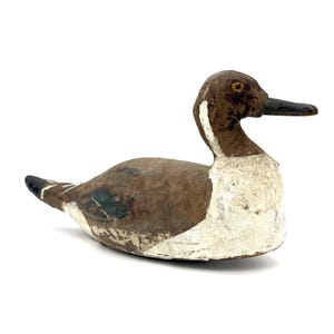 May include: A carved wooden duck decoy featuring a weathered brown head and back, a white chest, and a dark bill. The body displays patches of worn paint, including hints of green on the wings and a black tail tip, with a small orange eye detail.