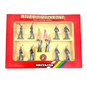 May include: A boxed set of hand-painted US Marine Corps model figures by Britains. The set includes ten miniature soldiers in dark blue uniforms with white hats, two carrying flags, displayed in a red box with clear plastic window packaging.