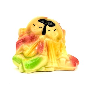 Puede incluir: Figura de cerámica que representa dos figuras sentadas. La figura presenta una paleta de colores amarillo, rojo y verde. Las figuras tienen detalles faciales negros. El diseño es una representación estilizada de un tema tradicional.