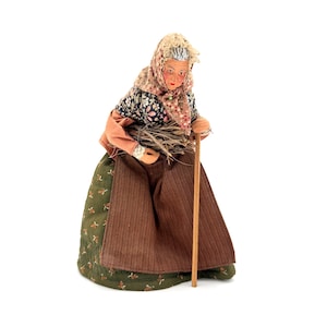 May include: A figurine of an elderly woman, dressed in a floral blouse, a patterned headscarf, and a long brown skirt. She carries a bundle of sticks in one arm and leans on a wooden cane. The figurine has a detailed, aged face.