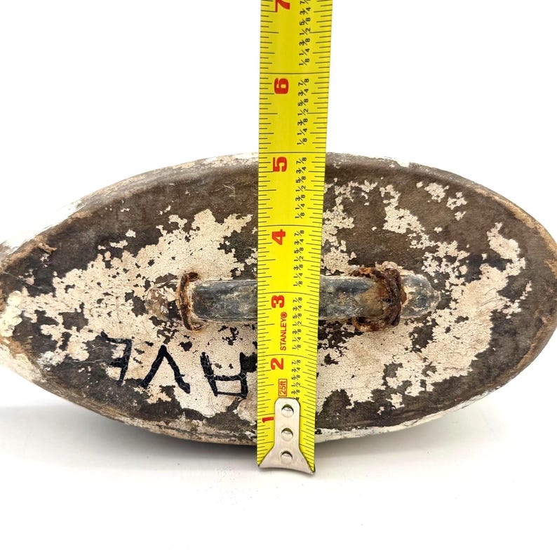 May include: The underside of a weathered wooden duck decoy featuring a dark brown and white distressed paint finish. A yellow Stanley measuring tape is positioned vertically across the center, showing a length of approximately 7 inches.