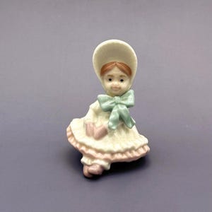 May include: A porcelain figurine of a seated girl wearing a bonnet and dress. The figurine has a light green bow, a pink purse, and a ruffled skirt with pink trim. The girl's hair is reddish-brown, and the figurine is on a grey background.