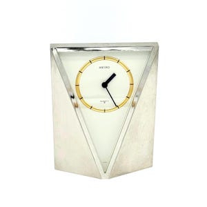 May include: A silver-toned Seiko quartz clock with a triangular face. The clock face is white with gold-coloured hands and a gold-coloured ring around the numbers. The word "SEIKO" is printed at the top of the clock face.