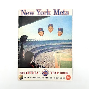 May include: A vintage 1969 New York Mets Official Year Book. The cover features the team's name in blue and white, with floating player heads above a stadium scene. The book includes the Shea Stadium location and a 50¢ price tag.