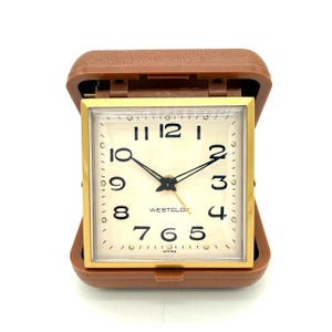 Könnte beinhalten: Ein Vintage-Reisewecker von Westclox in einem braunen Gehäuse. Das Zifferblatt ist quadratisch mit einem goldfarbenen Rahmen, schwarzen Ziffern und schwarzen Zeigern. Die Uhr ist geöffnet und zeigt das cremefarbene Zifferblatt mit dem Westclox-Logo.
