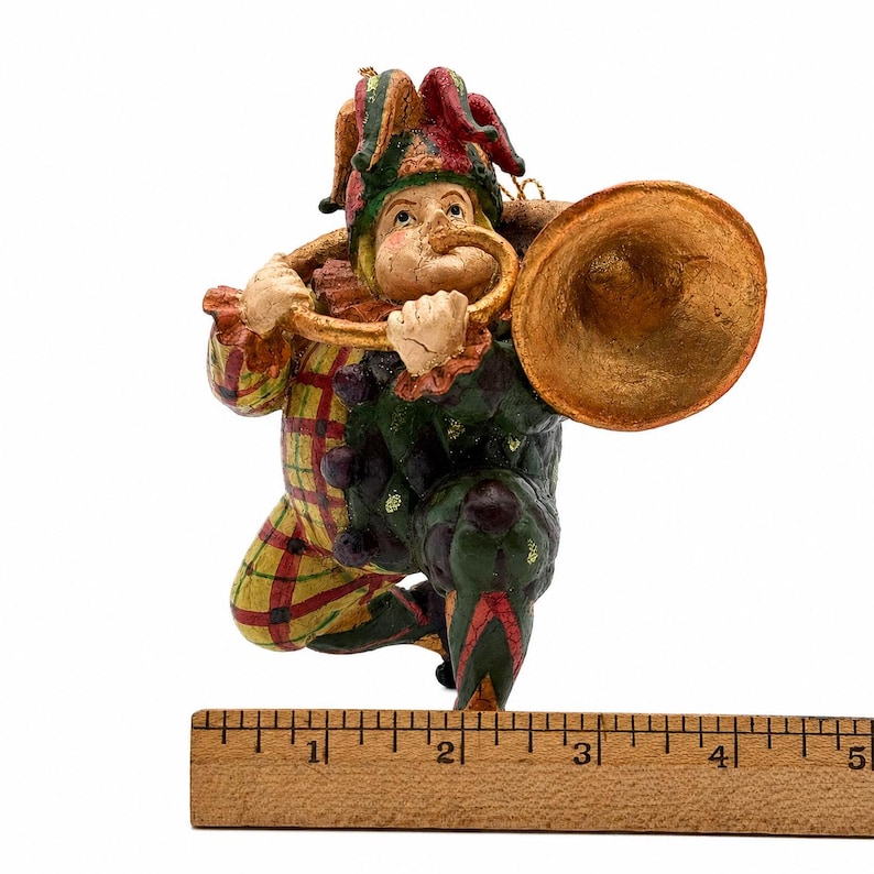 May include: A jester figurine wearing a red, yellow, and green checkered outfit and a matching hat, playing a large gold trumpet. The figure is positioned above a wooden ruler, showing a total width of approximately 5 inches from end to end.