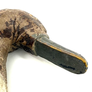 May include: A weathered, hand-carved wooden duck decoy head featuring a textured, brown-painted surface with visible signs of age and wear. The bill is painted a dark, muted gray with a thin yellow trim along the edge, showing a realistic form.