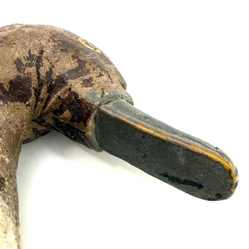 May include: A weathered, hand-carved wooden duck decoy head featuring a textured, brown-painted surface with visible signs of age and wear. The bill is painted a dark, muted gray with a thin yellow trim along the edge, showing a realistic form.