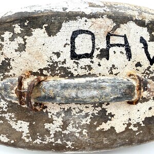 May include: A weathered, oval-shaped object with a dark, textured surface covered in cracked white paint. A horizontal metal handle with visible rust and two circular metal rings is attached to the center. The black letters DA are visible.