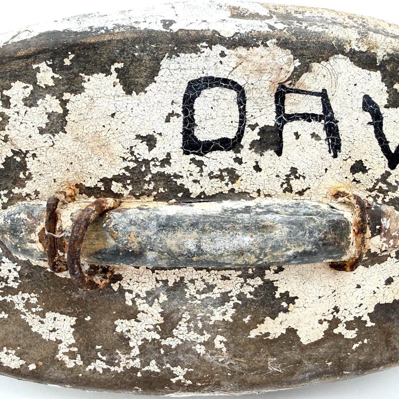 May include: A weathered, oval-shaped object with a dark, textured surface covered in cracked white paint. A horizontal metal handle with visible rust and two circular metal rings is attached to the center. The black letters DA are visible.