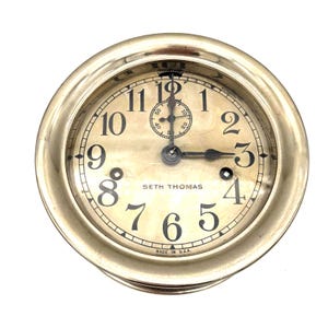 May include: A vintage Seth Thomas clock with a gold-toned casing and a cream-coloured face. Black numerals indicate the hours, with a small dial at the top. The clock hands show approximately 3:15. The words "SETH THOMAS" and "MADE IN U.S.A." are visible.