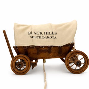 May include: A wooden covered wagon lamp featuring a dark brown stained wood frame and four spoked wheels. The beige fabric cover is printed with the black text "BLACK HILLS SOUTH DAKOTA". A white electrical cord extends from the center base.