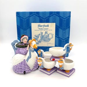 Puede incluir: Un juego de té "Mother Goose" de cerámica, que incluye una tetera y cuatro tazas. La tetera tiene forma de mujer con un vestido morado y un ganso. Las tazas son blancas y con forma de ganso, colocadas sobre bases con forma de libro morado. El juego viene en una caja azul.