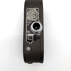 May include: A vintage Keystone Model K-8 8mm camera. The camera is black with a silver lens and a detailed information panel. The panel includes exposure settings for various lighting conditions, such as brilliant sunlight and hazy open scenes. The camera is marked "Made in U.S.A."
