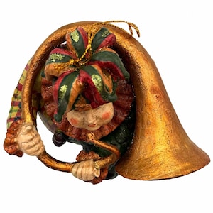 May include: A decorative ornament featuring a jester figure wearing a multi-colored hat with red and green segments and gold glitter accents. The jester holds a large, curved, metallic gold horn instrument and wears a ruffled collar.