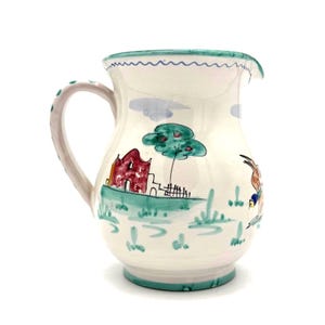 Könnte beinhalten: Weißer Keramikkrug mit handgemalter Landschaftsszene. Der Krug zeigt ein rotes Gebäude, einen grünen Baum und eine Figur mit einem Esel. Der Griff ist hellrosa mit grünen Punkten. Der Rand hat ein wellenförmiges blaues Design.