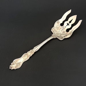 May include: Silver serving fork with ornate grape and leaf design. The fork features three tines and a decorative handle. The design includes cut-out details and a detailed grape cluster pattern. The serving fork is likely made of sterling silver.