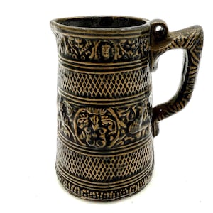 Puede incluir: Una jarra de cerámica marrón con asa. La jarra presenta intrincados patrones en relieve, incluyendo formas de diamante y diseños florales. El asa también está decorada con detalles tallados. La estética general es rústica y vintage.