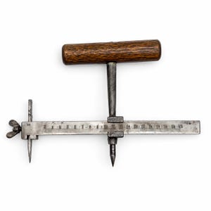 May include: A metal circle cutter tool featuring a wooden T-handle and a horizontal bar with a scale marked from 2 to 20 centimeters. The device includes an adjustable sliding center point and a cutting blade secured by a metal wing nut.