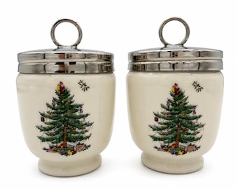 May include: Two cream-colored ceramic canisters with silver-toned metal lids featuring a circular handle. Each canister is decorated with a central illustration of a decorated Christmas tree with gifts at the base and a small Santa figure on top.