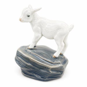 May include: A white ceramic goat figurine on a grey rock. The goat has a detailed texture and is posed with its head turned slightly. The rock has a glossy finish. A decorative item.